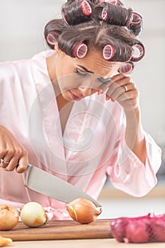 Woman in hair curlers and nighgown cries from onion cutting in the kitchen, wiping the tear from her eye