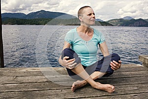 A woman with hair loss from chemotherapy treatment sits outside on a dock with the ocean and mountains behind her