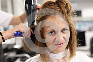 Woman hairdresser dries and straightens client hair with comb and hair dryer in beauty salon