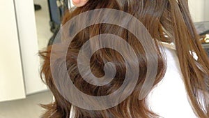 Woman hairdresser marcels long brown hair with curling iron in light hairdressing salon backside view