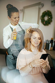 woman hairdresser in modern beauty salon with hair oil bottle and client with smartphone