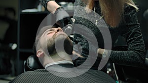 Woman hairdresser works with trimmer with a beard of a client.