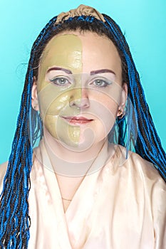 A woman with half of her face in a mask of green clay on a blue background