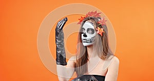 Woman with Halloween makeup with scary makeup adjusting her witch clothing long gloves over orange background.