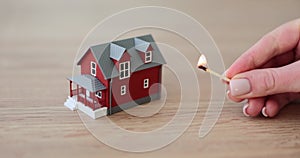 Woman hand brings lit match to house model on wooden table