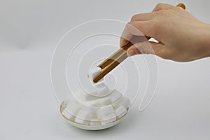 A woman hand clip a piece of sugar with bamboo clip, white cubes rock sugar on a plate, isolated on white background, diabetes