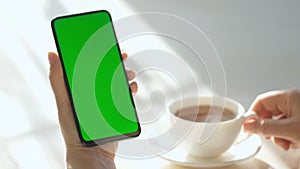 Woman Hand Cup of Hot tea and Using Smartphone Watching Green Screen Top View. Smartphone with Green Mock-up Screen