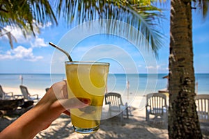 Woman hand with fresh juice on tropical beach landscape. Tropical island vacation on luxury resort