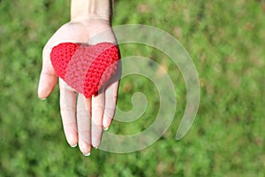 Woman hand giving red hand-made crocheted heart with green grass background and copy space. Valentine Day. Symbol of love.