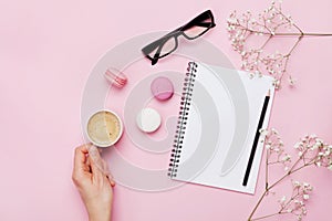 Woman hand hold cup of coffee, cake macaron, clean notebook, eyeglasses and flower on pink table from above. Female working desk.