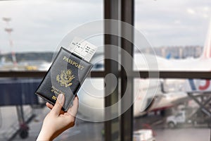 Woman hand holding american passport with boarding pass in the airport waiting zone looking through the window at planes.