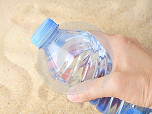 Woman hand holding a blue plastic bottle with drinking water over a sea sand. Concept of thirst, hot summer weather, sea holiday