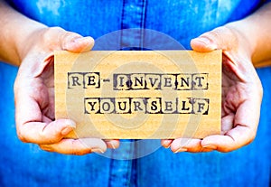 Woman hand holding cardboard card with words Re-Invent Yourself