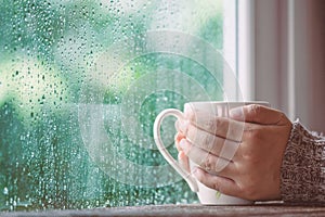 Woman hand holding the cup of coffee or tea on rainy day window