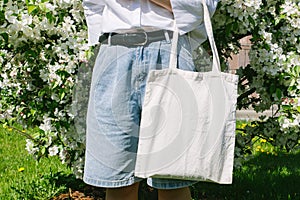 Woman hand holding eco bag. Female hold white canvas textille mesh zero waste shopper on city street, green apple tree