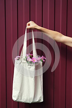 Woman hand holding eco reusable blank canvas shopping bag with flowers inside on red background. Zero waste mock-up. Vertical