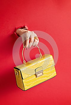 Woman hand holding fashion handbag through the hole in red paper background. Chic, style, fashion collection, trends, beauty blog