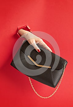 Woman hand holding fashion handbag through the hole in red paper background. Chic, style, fashion collection, trends, beauty blog