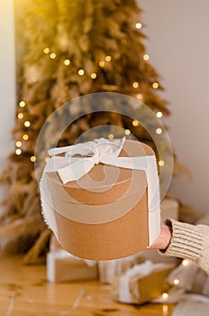 Woman hand holding gift New Year cardboard box with white bow, place for text or logo, with Christmas tree, garlands