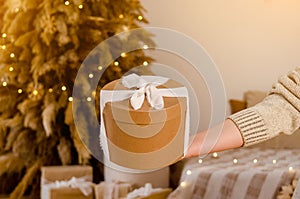 Woman hand holding gift New Year cardboard box with white bow, place for text or logo, with Christmas tree, garlands