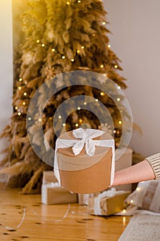 Woman hand holding gift New Year cardboard box with white bow, place for text or logo, with Christmas tree, garlands