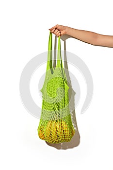 Woman hand holding green lime string shopping bag with vegetables and fruits in front of white wall. Eco lifestyle