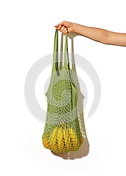 Woman hand holding green lime string shopping bag with vegetables and fruits in front of white wall. Eco lifestyle