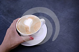 Woman hand holding latte coffee with white milk foam in heart shape on black background table