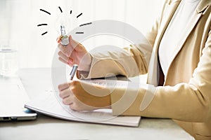 Woman hand holding light bulb, notebook, laptop, pen, documents on cream background with copy space. Creative idea, new