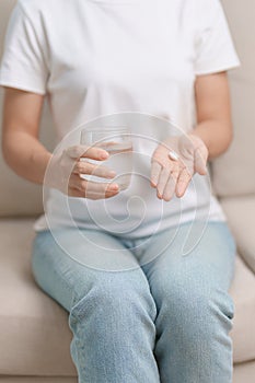 woman hand holding medicine painkiller pill and water glass on the sofa at home, taking for headaches, stomach ache, Diarrhea