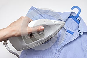 Woman hand holding a modern electrical white iron, a blue shirt on background, close up view - ironing, laundry and housework