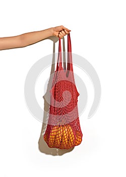 Woman hand holding red string shopping bag with vegetables and fruits in front of white wall. Eco lifestyle