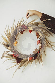 Woman hand holding rustic wreath on white wall background. Stylish boho autumn wreath with dried grass, berries, herbs and leaves