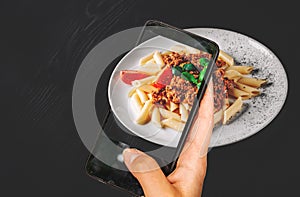 Woman hand holding and showing smart phone takes a photo Pasta penne bolognese in white plate on wooden table background