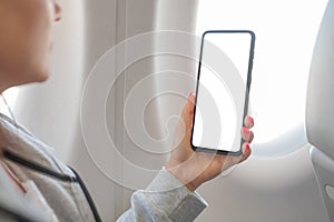 Woman hand holding smart cell phone with blank white screen on board of airplane near window seat. woman while flying on an