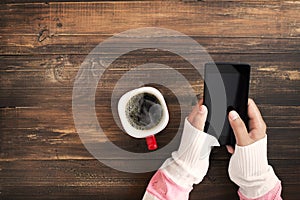 Woman hand holding smart phone with hot cup of coffee on wood table