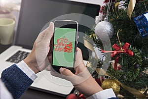 Woman hand holding smart phone with merry christmas text on desk