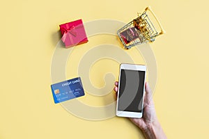 Woman hand holding smartphone, miniature of gift boxes in trolley and credit card on yellow background. Online shopping concept.