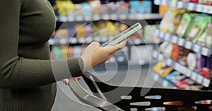 Woman hand holding smartphone and srolling screen in supermarket. Girl using phone while walking and pushing shopping