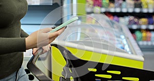 Woman hand holding smartphone and srolling screen in supermarket. Girl using phone while walking and pushing shopping