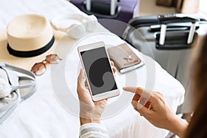 Woman hand holding smartphone with travel accessories on bed in hotel room. Travel, relaxation, journey, trip and vacation