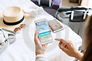 Woman hand holding smartphone with travel accessories on bed in hotel room. Travel, relaxation, journey, trip and vacation
