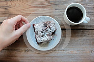 Woman hand holding a source with piece of chocolate cake. Cup of coffee and handmade brownie with cherries on a white plate