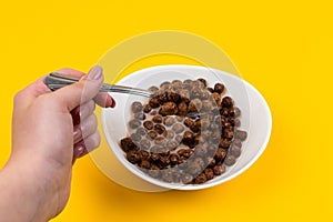 Woman hand holding spoon at White bowl with chocolate corn cereal balls and milk on yellow background, top-down view. Modern fresh