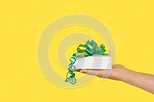 Woman hand holding a white gift box with a green ribbon on a yellow background