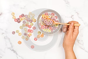 Woman hand holding a wooden spoon on bowl with fruit flavor colored cereals with milk on a kitchen marble counter