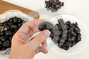 Woman hand holds black olive crushed with a knife for pitting it on a background of kithen table with plate and chopping board.