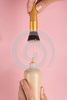 Woman hand holds a bottle of foundation, puts on a brush, on a pink background. Makeup artist work concept, copy space