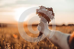 Woman hand holds a bouquet of wildflowers against a sunset background