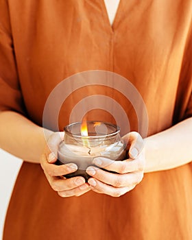 Woman hand holds lit scented candle in glass jar with natural ingredients on terracotta